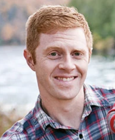 A man with short red hair and fair skin smiles at the camera. He is wearing a red and blue plaid shirt, and the background shows a blurred outdoor scene with water and trees.